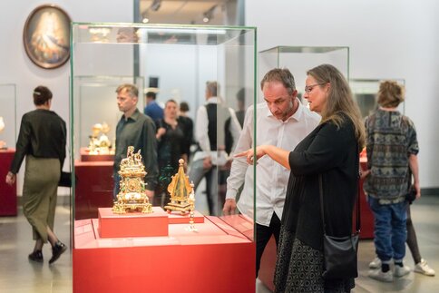 Zwei Personen betrachten Ausstellungsstücke in einer Vitrine, im Hintergrund sind weitere Personen sowie Vitrinen sichtbar