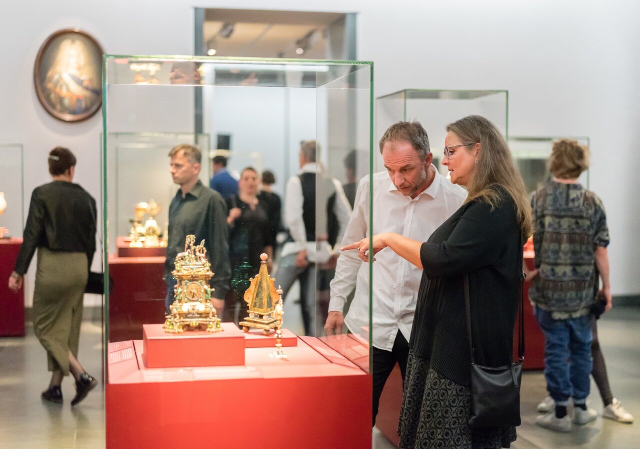 Zwei Personen betrachten Ausstellungsstücke in einer Vitrine, im Hintergrund sind weitere Personen sowie Vitrinen sichtbar